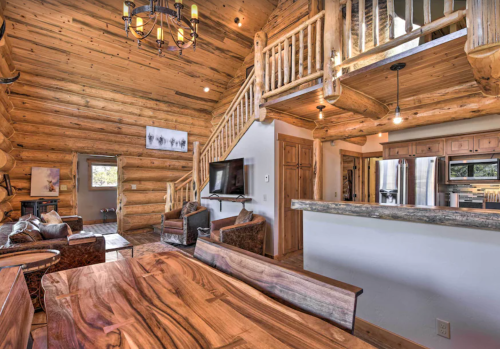 Cozy log cabin interior featuring a wooden dining table, living area, and staircase with rustic decor and warm lighting.