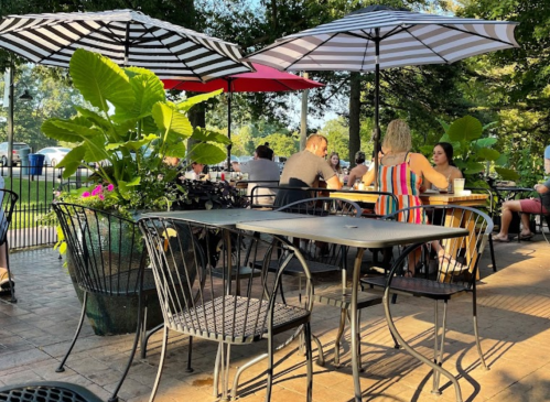 Outdoor dining area with tables, umbrellas, and plants, filled with people enjoying a sunny day.