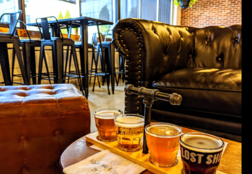 A wooden flight of four craft beers on a table, with a cozy leather couch and bar stools in the background.