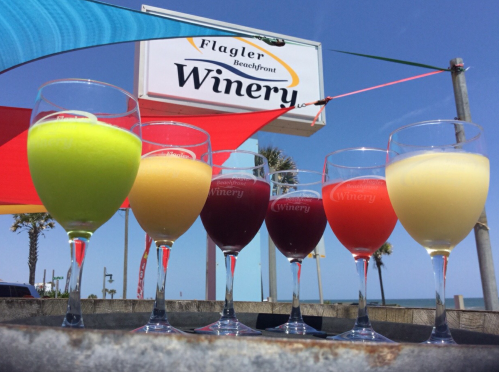 Six colorful wine glasses in front of a sign for Flagler Beachfront Winery, with a clear blue sky in the background.