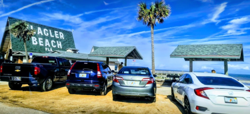 Cars parked in front of a beach building with a sign reading "Flagler Beach, FL" under a blue sky with palm trees.