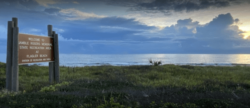 Sign welcoming visitors to Gamble Rogers Memorial State Recreation Area at Flagler Beach, with ocean and cloudy sky in background.