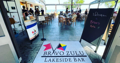Entrance to Bravo Zulu Lakeside Bar, featuring outdoor seating and a welcoming sign. Scenic view in the background.