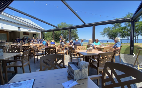 A lively outdoor dining area with wooden tables, people enjoying meals, and a view of the lake on a sunny day.