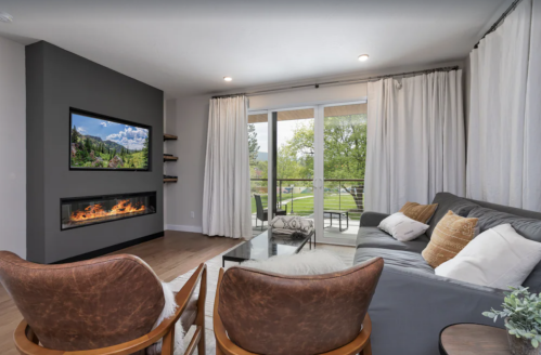 Modern living room with a gray sofa, two brown chairs, a wall-mounted TV, and a fireplace, featuring large windows.