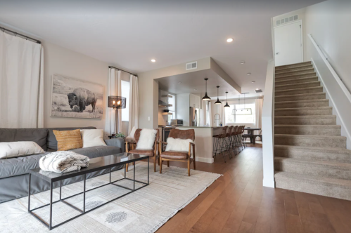 Modern living room with a gray sofa, wooden chairs, and a staircase leading to an upper level. Bright and inviting space.