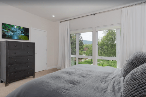 A cozy bedroom featuring a gray bed, dresser, and large windows with a view of greenery outside.