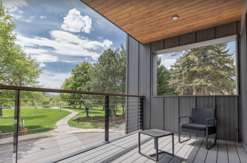 A modern balcony with a chair and table, overlooking a green park and blue sky with clouds.