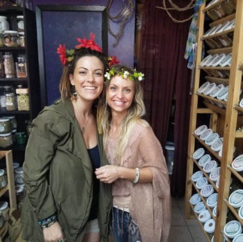 Two women with flower crowns smile together in a cozy shop filled with jars and herbs.