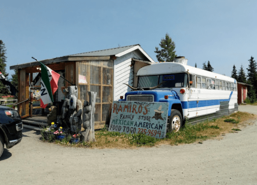 A blue and white bus parked outside a small building with a sign for Ramiro's Mexican American food and a Mexican flag.