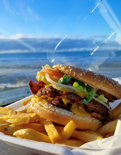 A juicy burger with bacon, lettuce, tomato, and cheese, served with golden fries against a scenic ocean view.