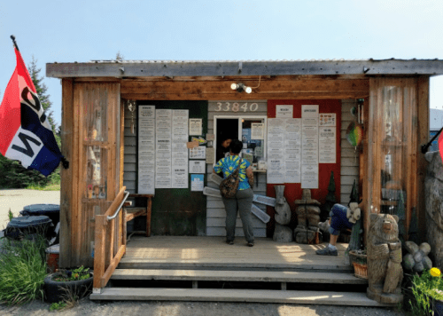 A person in a tie-dye shirt approaches a rustic wooden building with menus posted outside. Flags and sculptures decorate the entrance.