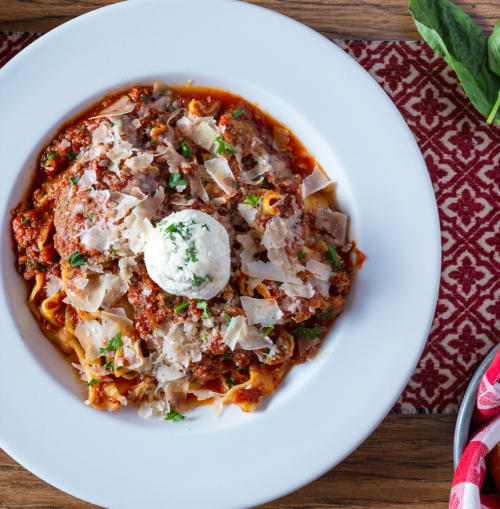 A plate of pasta topped with meatballs, marinara sauce, shaved cheese, and a dollop of creamy ricotta, garnished with herbs.