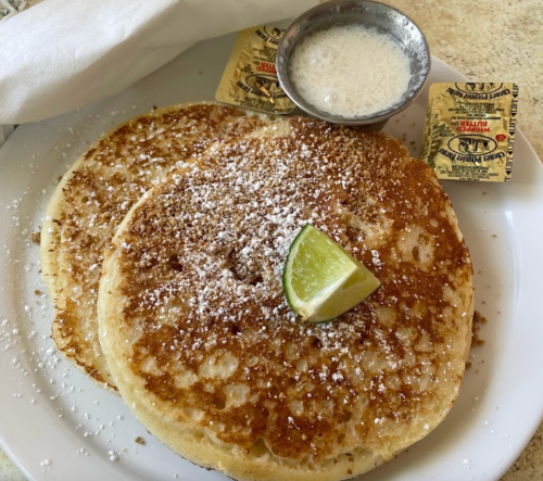 Two golden pancakes topped with powdered sugar and a lime wedge, served with butter and syrup on the side.
