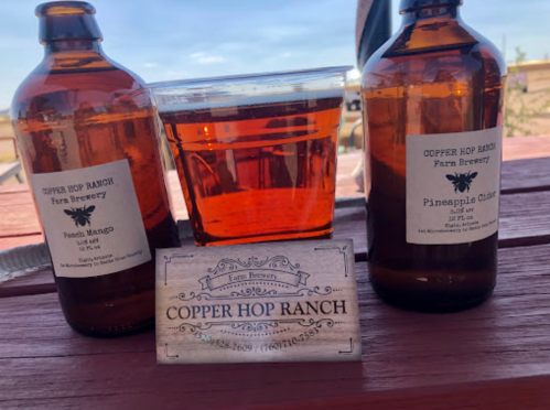 Two brown bottles labeled "French Mango" and "Pineapple Cider" beside a clear cup of beer and a business card on a table.