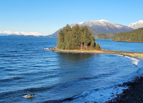 A serene coastal scene featuring a small island surrounded by blue water and snow-capped mountains in the background.
