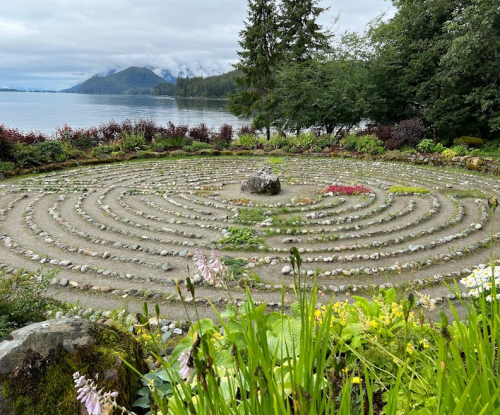 A serene stone labyrinth surrounded by lush greenery and a calm lake, with mountains in the background under a cloudy sky.