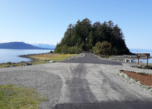 A paved path leads towards a forested area by the water, with mountains visible in the distance under a clear blue sky.