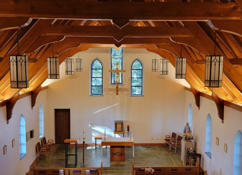 Interior of a church with wooden beams, stained glass windows, and an altar at the front, illuminated by natural light.