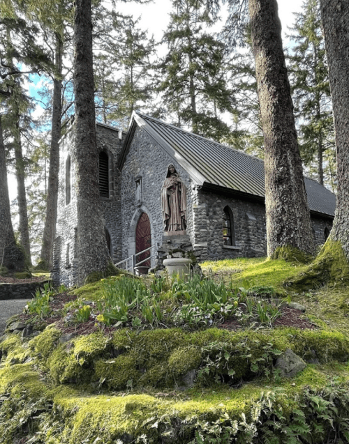 A stone church surrounded by tall trees and lush greenery, featuring a statue in the foreground.