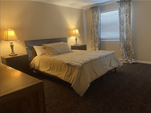Cozy bedroom with a bed, nightstands, lamps, and patterned curtains, featuring warm lighting and a soft carpet.