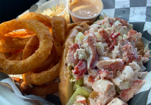 A close-up of a lobster roll with lettuce, served with onion rings and a small cup of sauce on the side.