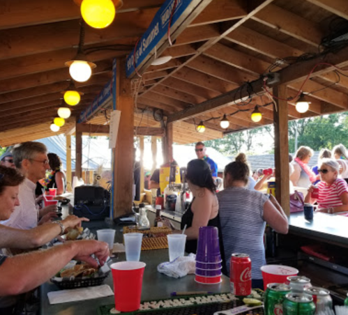 A lively outdoor bar scene with patrons enjoying drinks and food under a wooden roof with string lights.