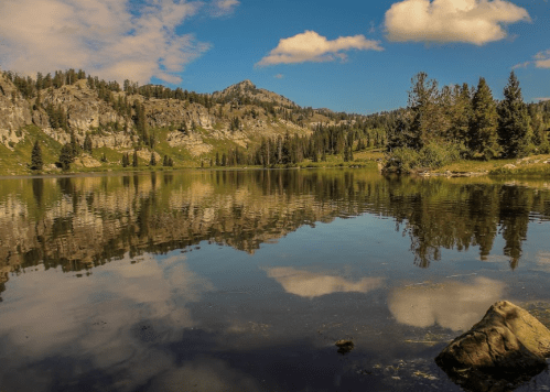 A serene lake reflecting mountains and clouds, surrounded by lush greenery and trees under a blue sky.