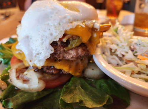 A stacked burger with a fried egg, cheese, and vegetables, served with a side of coleslaw on a lettuce leaf.
