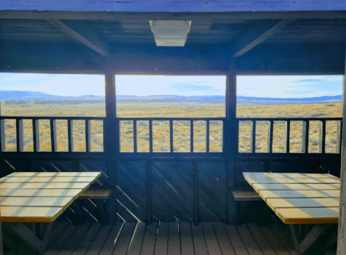View from a wooden shelter with two tables, overlooking a vast, open landscape and distant mountains.