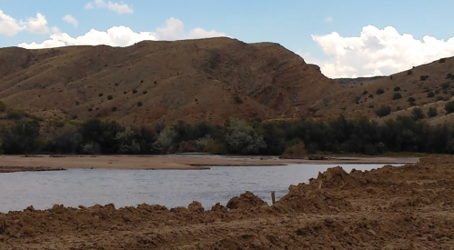 A serene river flows through a sandy landscape, surrounded by hills and scattered trees under a partly cloudy sky.
