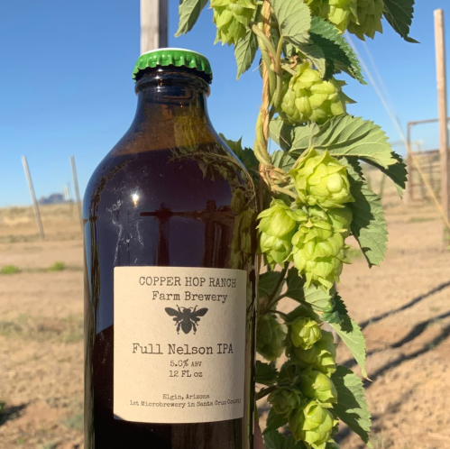 A bottle of Full Nelson IPA from Copper Hop Ranch, with hops growing nearby in a sunny field.