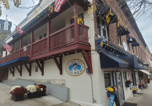 Exterior of Salmon Run Fish House, featuring a red balcony, blue awnings, and seasonal flower planters.