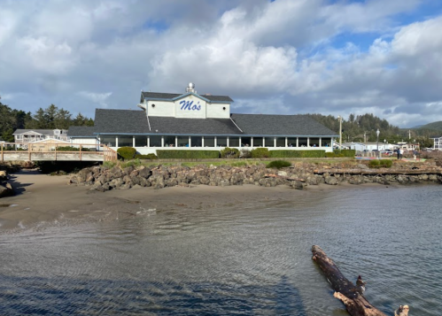 A waterfront restaurant with a blue sign reading "Mo's," surrounded by rocks and a sandy beach under a cloudy sky.