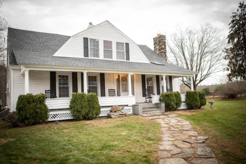 A charming white house with a gray roof, front porch, and stone pathway, surrounded by greenery and trees.
