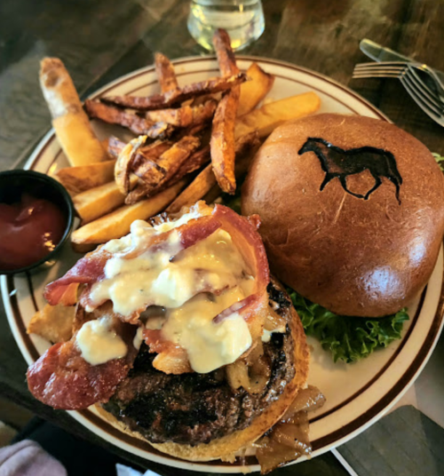 A gourmet burger topped with bacon and sauce, served with fries and a bun featuring a horse logo.