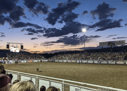 A rodeo event at dusk, with a crowd watching and a large screen displaying the action under a colorful sky.