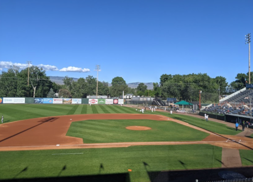 A sunny baseball field with a well-maintained diamond, surrounded by trees and a seating area in the background.
