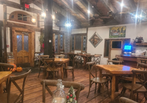 Cozy restaurant interior with wooden tables, chairs, and rustic decor, featuring a bar and warm lighting.