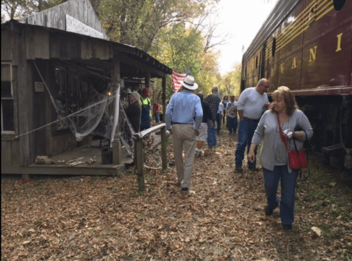 A crowd walks along a path beside a vintage train and an old wooden building, surrounded by autumn leaves.