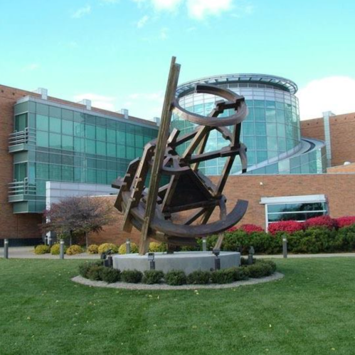 Abstract metal sculpture in a grassy area, with a modern glass building in the background and colorful shrubs.