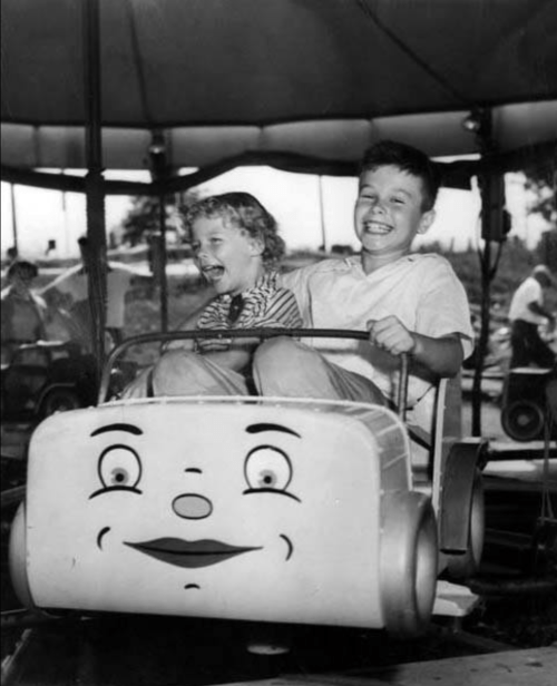 Two children joyfully ride a vintage amusement park ride, smiling and laughing under a carousel canopy.