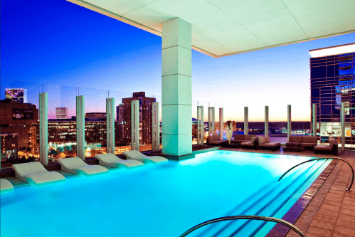 Rooftop pool with lounge chairs, overlooking a city skyline at sunset, with vibrant blue and purple hues in the sky.