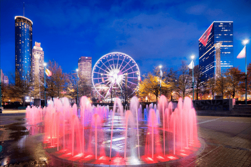 A vibrant cityscape at night featuring a colorful fountain, a Ferris wheel, and illuminated skyscrapers.