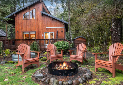 Cozy outdoor seating area with four chairs around a fire pit, set against a wooden cabin and lush greenery.