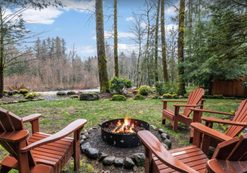Cozy outdoor seating around a fire pit, surrounded by trees and a river in a serene natural setting.