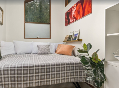 Cozy room with a bed covered in a plaid blanket, decorative pillows, and plants, featuring large windows and artwork.