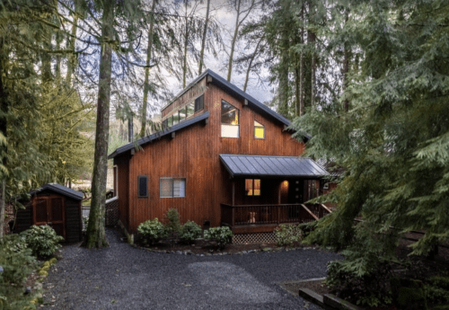 A wooden cabin surrounded by tall trees, featuring a sloped roof and a gravel pathway leading to the entrance.