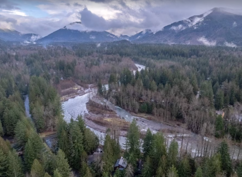 Aerial view of a winding river surrounded by dense forests and mountains under a cloudy sky.