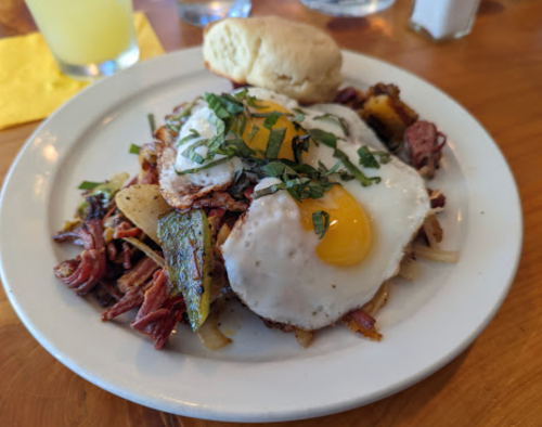 A plate of hash with corned beef, potatoes, two fried eggs, and a biscuit, garnished with fresh herbs.